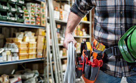 Person wearing a tool belt with various hand tools shopping in a hardware store aisle stocked with supplies