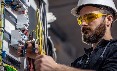Electrician wearing a white hard hat and yellow safety glasses working on electrical wiring in an industrial control panel